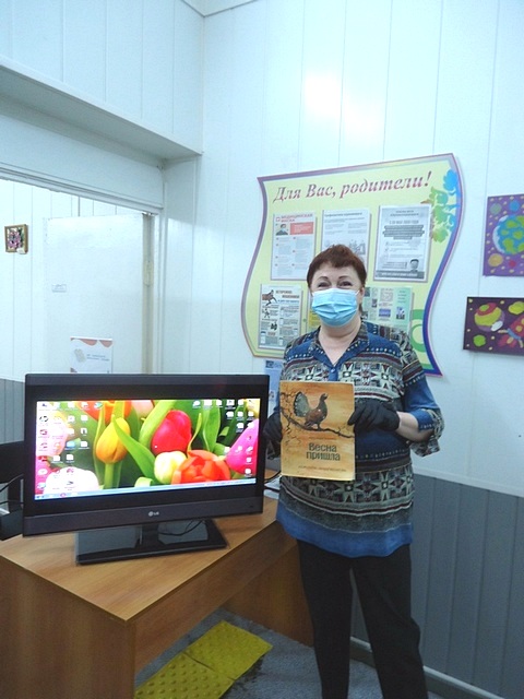 Перед экраном с изображением цветов стоит бибилотекраь и держит в руке детскую  книгу 